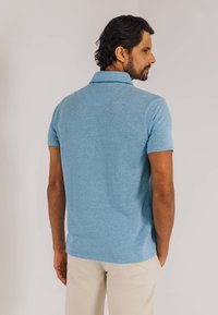 Lichtblauw poloshirt met korte mouwen, fijne textuur, kraag en een regular fit, gecombineerd met beige broek. Gezien van de achterkant.