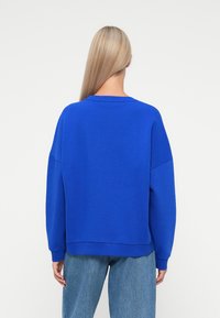 Hanorac oversized albastru cu decolteu rotund, umeri căzuți, manșete și tiv din tricot; asortat cu blugi albastru deschis.
