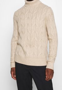 Suéter de punto beige con cuello alto y puños y dobladillo acanalados. Presenta un patrón texturizado y un diseño ajustado. Se lleva sobre pantalones oscuros.