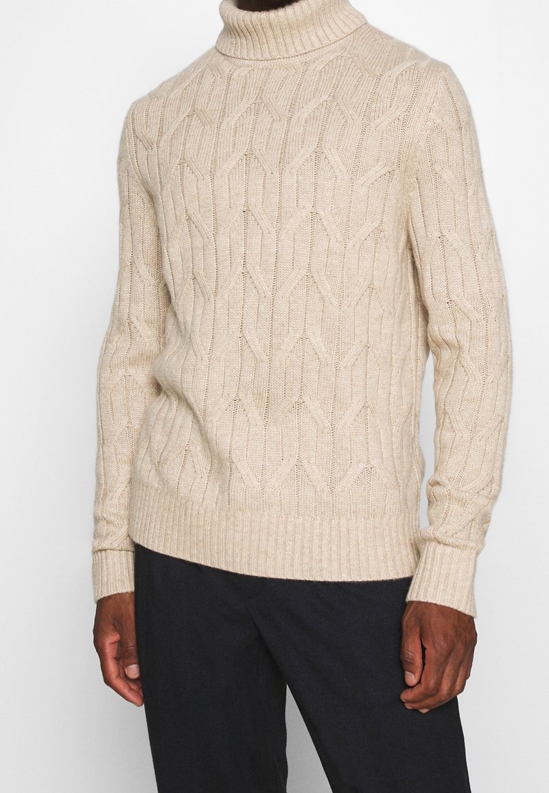 Suéter de punto beige con cuello alto y puños y dobladillo acanalados. Presenta un patrón texturizado y un diseño ajustado. Se lleva sobre pantalones oscuros.