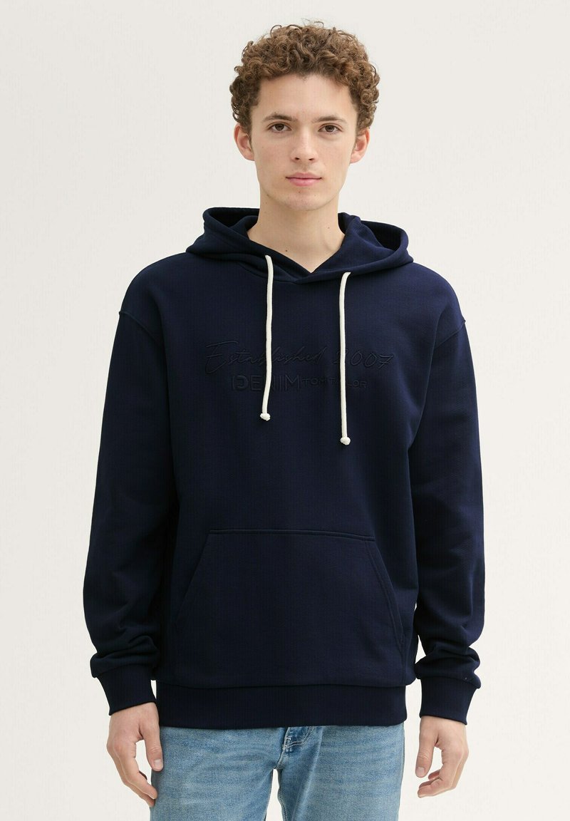 Dunkelblaue Kapuzenjacke aus strukturiertem Stoff, mit Kordelzugverschluss und einer frontalen Kängurutasche. Mit geprägtem Logodesign.