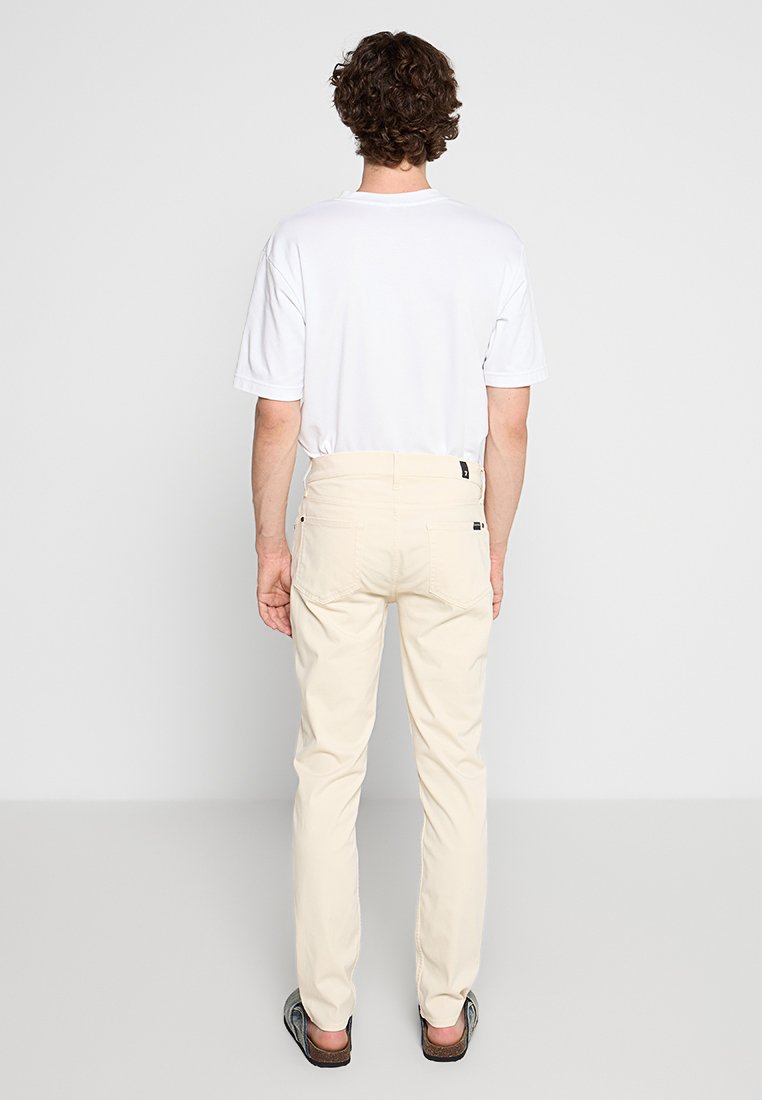 Beige katoenen broeken met een rechte pijp, voorzien van achterzakken en een logo label op de tailleband, gecombineerd met een plain witte t-shirt.