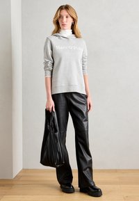 Grauer Kapuzenpullover mit weißem Logo, kombiniert mit schwarzen Kunstlederhosen. Das Modell hält eine schwarze gerippte Tragetasche und steht auf Holzboden.