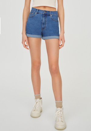 Kvinde iført opsmøgede blå cowboyshorts, beige ankelsokker og hvide høje sneakers, stående mod en ensfarvet baggrund.