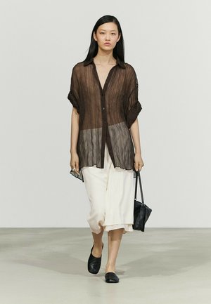 Jeune femme marchant avec une chemise marron foncé transparente à boutons, une jupe blanche ample, des chaussures noires sans lacets, tenant un petit sac à main noir dans la main droite.