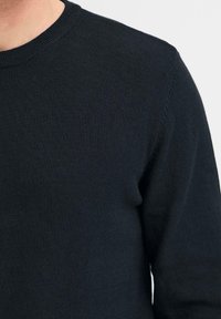 Primo piano di una persona che indossa un maglione nero a maniche lunghe con scollo rotondo su uno sfondo chiaro e uniforme.
