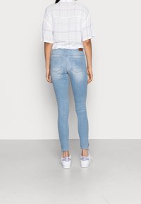 Persona de pie de espaldas, vistiendo jeans ajustados de color azul claro, zapatillas blancas con acentos morados y una camiseta blanca de manga corta con un patrón de cuadrícula.