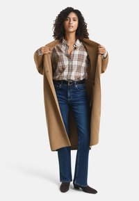 Camelkleurige jas, geruite shirt in bruin en crèmekleuren, donkerblauwe jeans, donkergroene hakken, gepresenteerd in een staande pose met open armen.
