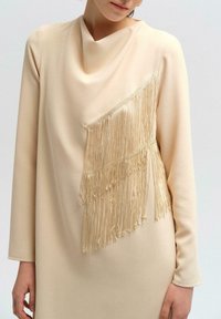 Lichtbeige jurk met een kraag en een diagonale gouden fringe-detail aan de voorkant. Lange mouwen van een gladde stof.