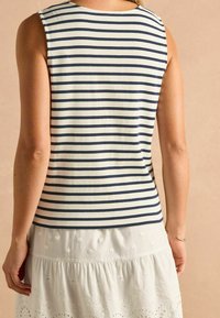 Top bez rukávov s námorníckymi a bielymi pruhmi, vyrobený z mäkkej tkaniny. Spodný lem má rovný strih. Nosený s textúrovanou bielou sukňou.