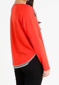 Camiseta roja de manga larga con cuello redondo, que presenta un dobladillo a rayas. Tejido suave con un ajuste holgado y un diseño en la parte posterior ligeramente curvado.