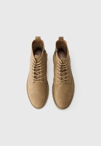Par de botas de ante de color marrón claro con cordones, cremalleras laterales, puntas redondeadas y lengüetas en un fondo liso gris claro.
