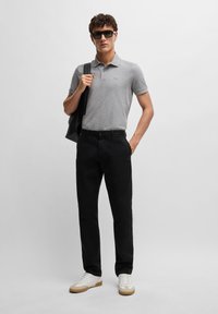 Pelēks polo krekls, melni bikses, baltas kedas ar brūniem akcentiem un saulesbrilles. Modelis tur melnu somu un stāv pret neitrālu fona.