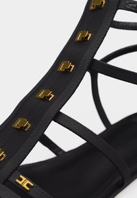 Μαύρο σανδάλι με λουριά και χρυσές τετράγωνες καρφίτσες κατά μήκος του κάθετου λουριού σε λευκό φόντο.