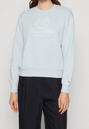 Kobieta ubrana w jasnoniebieską bluzę Tommy Hilfiger z logo i czarne spodnie z białymi paskami po bokach na tle jednolitego tła.