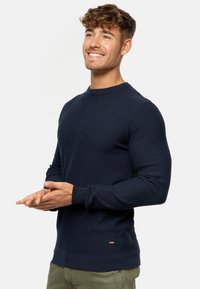 Marineblå strikket sweater med rund hals, ribbede manchetter og kant. Har et struktureret stof og en lille logo detalje nær taljen.