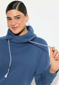 Pullover blu con colletto alto a scialle, caratterizzato da un cordoncino bianco e navy attorcigliato con estremità a frange. Tessuto morbido e vestibilità rilassata.