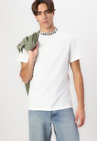 T-shirt en coton blanc avec un col noir et blanc haut, arborant un petit logo sur la poitrine. Le modèle tient un pull vert sur une épaule.
