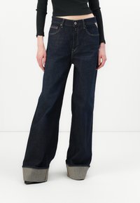 Wijde donkere denim jeans met een hoge taille. Voorzien van een knoopsluiting, zakken aan de voorkant en opgerolde manchetten met contrasterende stof.