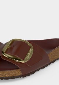 Καφέ σανδάλι Birkenstock με μεγάλο χρυσό αγκράφα και φερμουάρ φελλού, λοξή πλαϊνή όψη σε λευκό φόντο.
