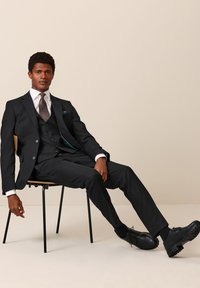 Abito nero con gilet abbinato, camicia bianca e cravatta a motivi. Scarpe in pelle nere con suole testurizzate. Seduto su una sedia minimalista.