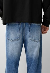 Svetlo modri kavbojke z vintage teksturo, dvema zadnjima žepoma in sproščenim krojem, v kombinaciji s črno majico z dolgimi rokavi.