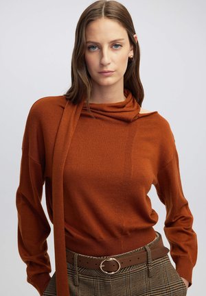 Pull en couleur rouille avec un col montant, des manches longues et un design texturé. Associé à un pantalon à motifs et une ceinture marron.