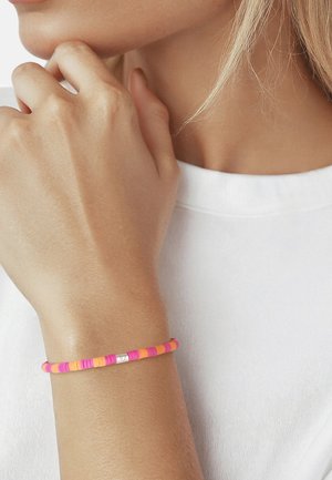 Femme portant une chemise blanche avec un bracelet perlé rose et orange au poignet, main reposant près du menton.