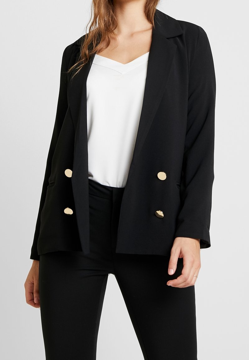 Femme portant un blazer noir croisé à boutons dorés sur un chemisier blanc à col en V et un pantalon noir, debout devant un fond uni.