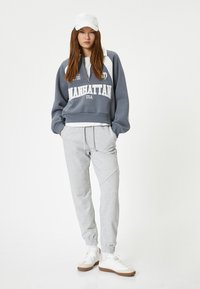 Felpa grigia con la scritta "MANHATTAN USA", abbinata a pantaloni jogger tapered grigi chiaro con polsini elastici e un cappellino grigio chiaro. Sneakers bianche.