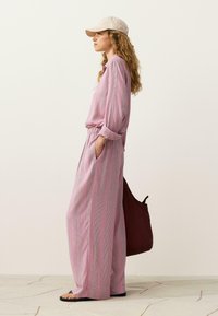 Femme portant une chemise ample à rayures roses et un pantalon avec les mains dans les poches, coiffée d'une casquette blanche et portant des sandales noires, tenant un grand sac foncé.