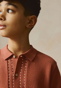 Polo a maglia colore ruggine con motivo a rilievo, chiusura con bottoni e dettaglio di ricamo bianco lungo la patta dei bottoni.