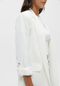 Blazer blanco con un diseño estructurado, que presenta un cuello con muesca, mangas enrolladas y detalles de botones. Hecho de una tela suave con un corte ajustado.