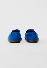 Ett par blåa slip-on skor med elastisk baksida och svart texturerad sula, sedd bakifrån mot en vit bakgrund.