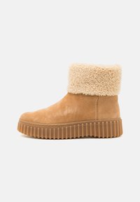 Botines de ante en tono arena con un suave puño de crema, suela de goma texturizada y detalles acanalados. Punta redondeada y costuras laterales.