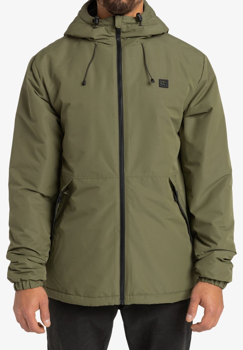 Giacca isolata verde oliva con cappuccio, chiusura con zip, polsini elasticizzati e tasche laterali. Presenta una patch con logo sul petto e una texture liscia.