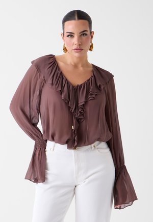 Žena u prozirnoj smeđoj bluzi s volanima i zvonolikim rukavima i bijelim hlačama, okrenuta prema naprijed na jednobijeloj pozadini.