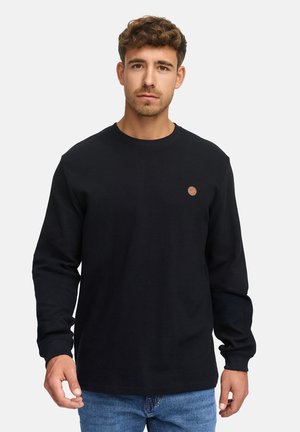 Schwarz langärmliges T-Shirt mit strukturiertem Rippstoff und rundem Halsausschnitt; auf der linken Brust befindet sich ein kleines braunes Logo. Kombiniert mit blauen Jeans.