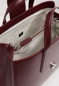 Bordeauxrode lederen handtas met twee handvatten, zilverkleurige hardware en een binnenzak met rits. De voering is van lichtgrijze stof.
