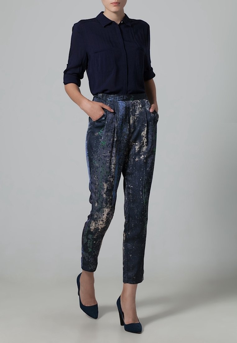 Femme portant une chemise bleu marine boutonnée, un pantalon fuselé à motif abstrait et des talons hauts bleu marine, debout, les mains dans les poches.