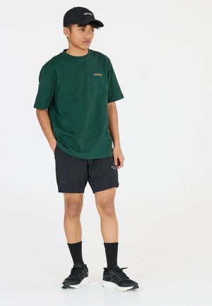 Grøn bomulds T-shirt med teksten "LES ARCS", parret med sorte shorts, sorte sokker og sorte sneakers. Mand står med hånden i lommen.