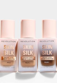 Tres botellas de Revolution Skin Silk Radiant Satin Serum Foundation. Cada una tiene un diseño mate en pastel rosa con letras doradas, con tres tonos visibles.