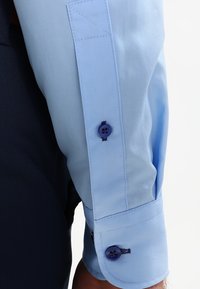 Lys blå skjorte med en glatt tekstur, med en knappet mansjett med en mørk blå knapp og sydd detaljer.