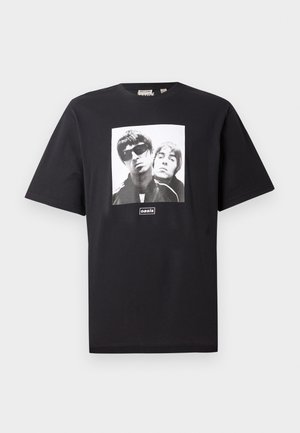 Must õhukese musta puuvilla t-särk, millel on ruudu kujuline valge graafika kahe figuuriga ja logo "oasis" pildi all. Lühikesed varrukad ja ümmargune kaelus.