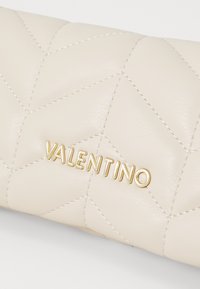 Prešívaná béžová kožená kabelka s zlatým nápisom "VALENTINO", doplnená jemným prešívaním a hladkou textúrou.