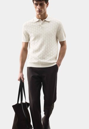 Uomo che indossa una polo a maniche corte bianca con motivo, pantaloni scuri, scarpe nere, che porta una grande borsa tote nera nella mano sinistra.