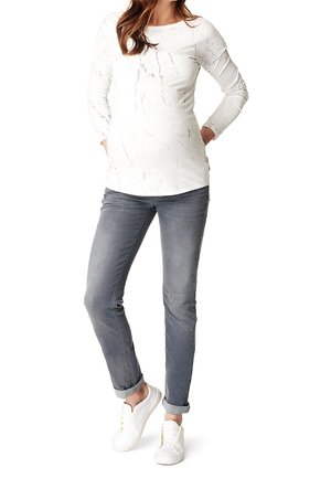 Frau steht und trägt ein weißes Langarmshirt mit dezentem Muster, graue hochgekrempelte Jeans und weiße Turnschuhe mit neongrünen Schnürsenkeln.