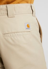 Beige cargo-housut, jotka on valmistettu kestävästä kankaasta ja joissa on takatasku sekä neliömainos Carhartt-logo oranssilla ja valkoisella.