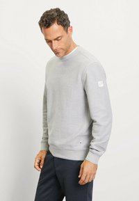 Grauer Rundhals-Pullover aus einem weichen Stoff mit gerippten Bündchen und Saum; hat ein kleines Logo-Patch am linken Ärmel. Getragen mit dunklen Hosen.