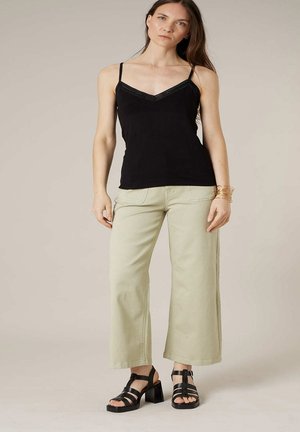 Femme debout portant un haut noir sans manches, un pantalon beige à jambes larges, des sandales à talons noires et des bracelets dorés au poignet gauche.